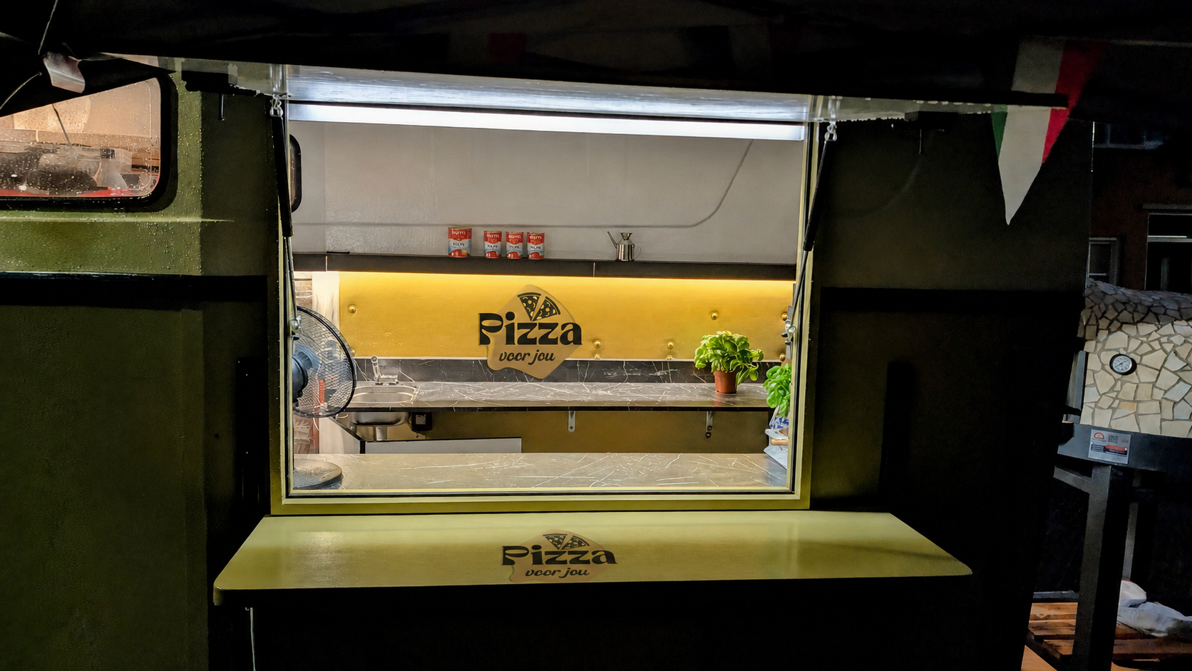 De pizzatrailer in de avond met verlicht uitgifteluik en logo