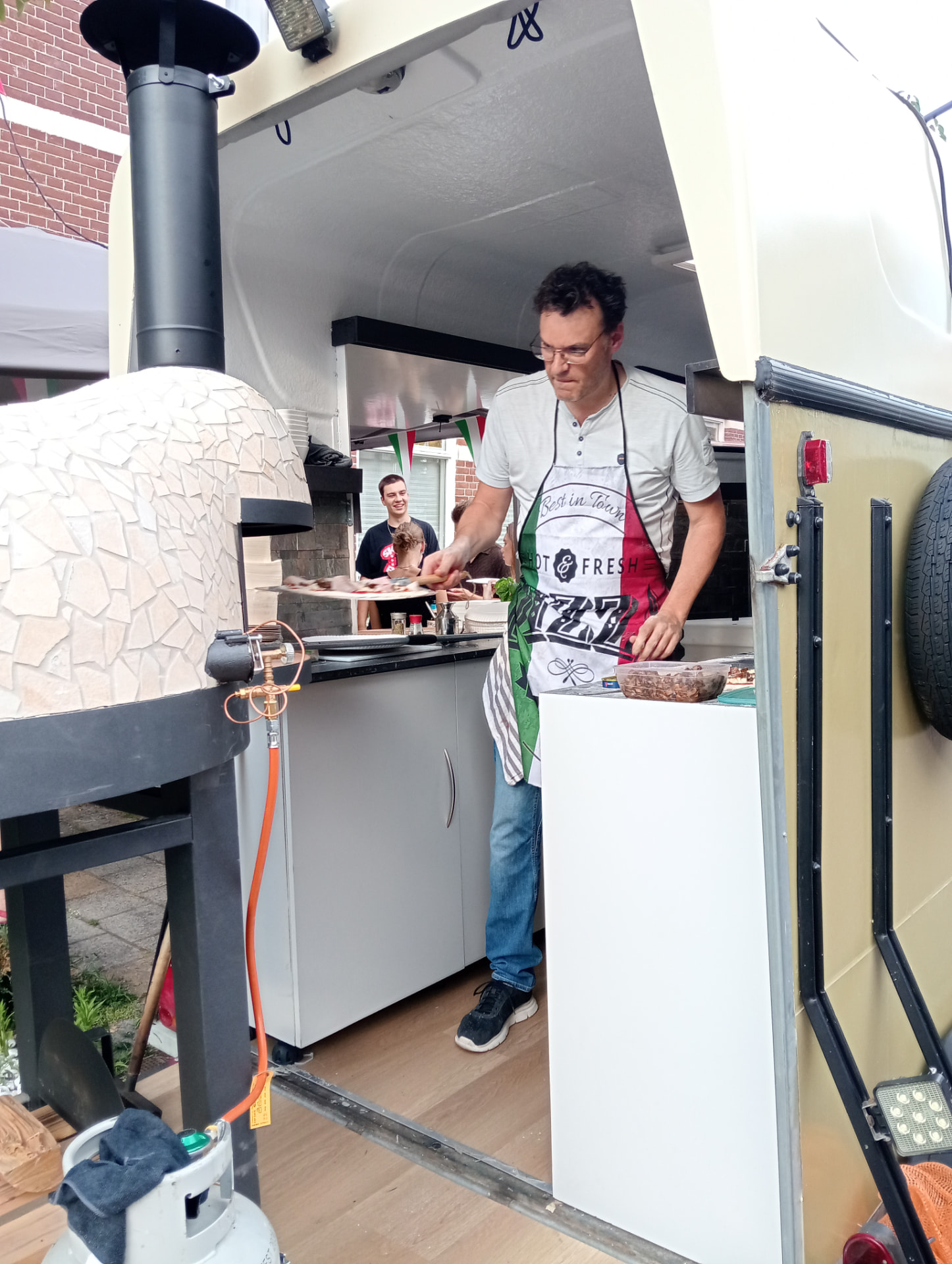 Frans schuift een pizza op de schep, gasten kijken toe
