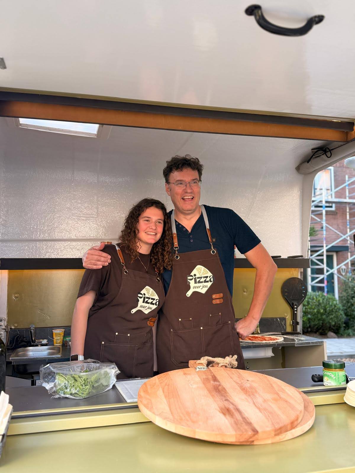 Twee mensen van Pizza voor jou glimlachen in het serverluik van de pizzatrailer in Gouda
